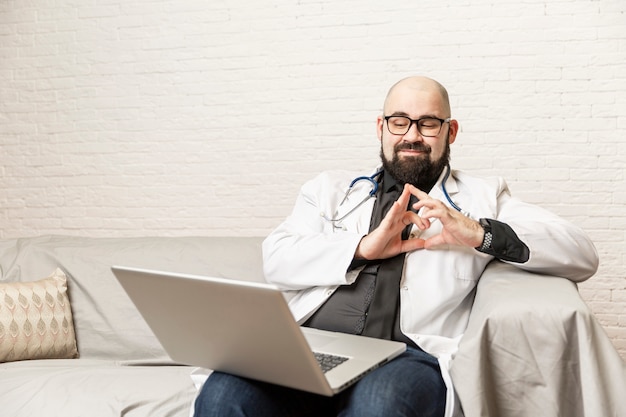 männlicher Arzt sitzt auf einem Sofa und arbeitet vor einem Laptop. Fernarbeit, Online-Konsultationen während der Epidemie.