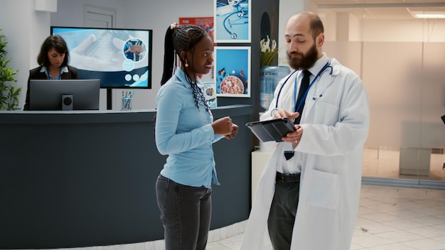 Männlicher Arzt spricht mit afroamerikanischen Patienten über Gesundheitsversorgung und zeigt medizinische Diagnoseergebnisse auf einem digitalen Tablet in der Wartezimmer-Lobby an der Krankenhausrezeption. Kontrollbesuch in der Einrichtung.