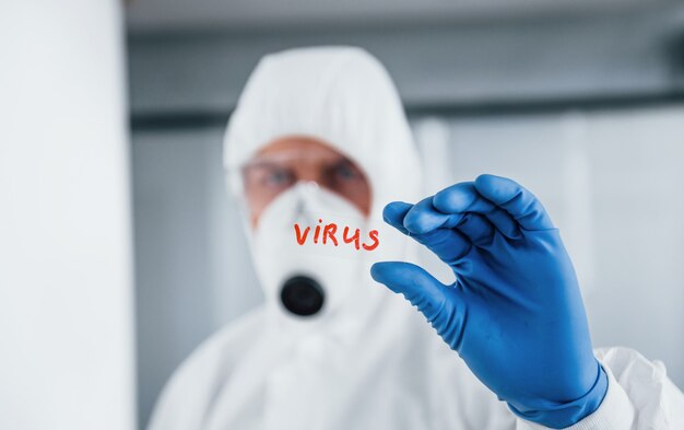 Foto männlicher arzt wissenschaftler im laborkittel, in der defensiven brille und in der maske hält glas mit viruswort darauf