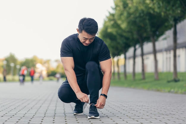 Männlicher asiatischer Athlet, der vor dem Joggen Schnürsenkel an einem Sneaker bindet und im Park in der Nähe des Stadions läuft