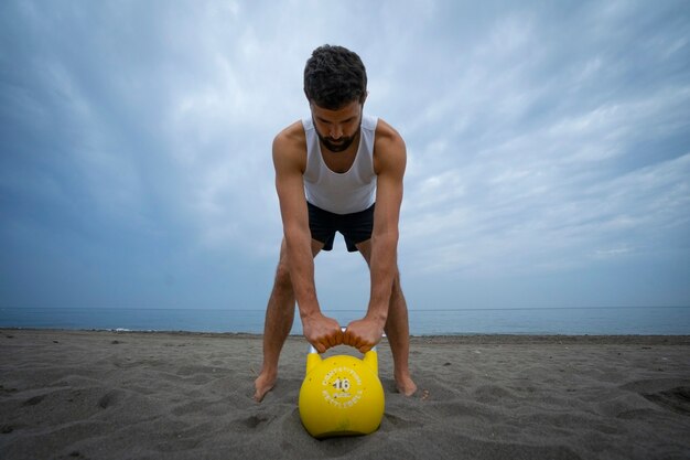 Männlicher Athlet, der am Strand mit Kesselglocken trainiert