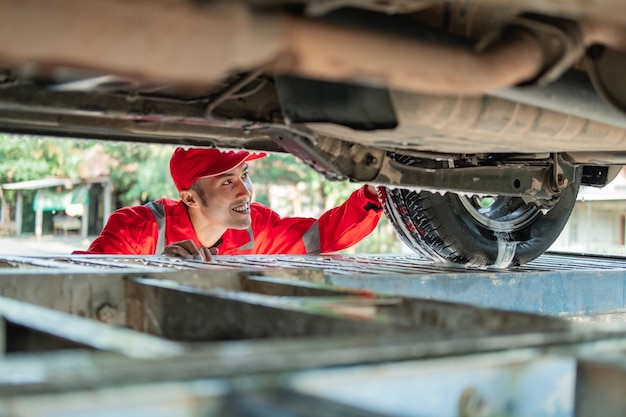 Männlicher Auto-Reiniger, der die rote Uniform trägt, schaut unter das Auto, während das Auto im Autosalon gewaschen wird