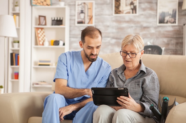 Männlicher bärtiger Arzt im Pflegeheim, der älteren Frauen hilft, ihren Tablet-Computer zu benutzen.