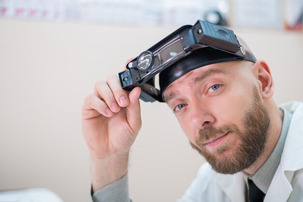 Männlicher bärtiger Arzt in einem medizinischen Mantel und mit einem Vergrößerungskopfband mit Lichtern, die auf dem Schreibtisch sitzen Optometrist-Ausrüstung Ärztliches Büro