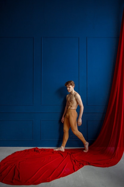 Männlicher Balletttänzer posiert an der blauen Wand im Tanzstudio, rotes Tuch. Performer mit muskulösem Körper, Anmut und Eleganz der Bewegungen