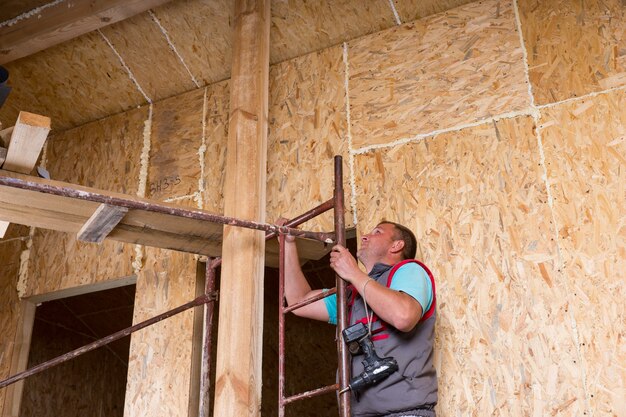 Männlicher Bauarbeiter Baumeister mit Akku-Bohrschrauber klettert Leiter von Gerüsten im unfertigen Haus mit freiliegendem Spansperrholz-Rahmen hoch