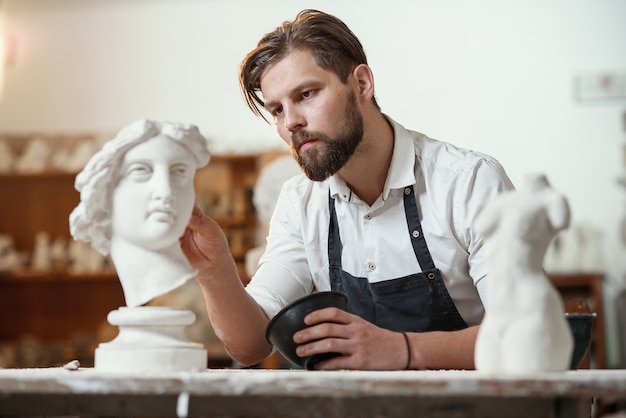 Männlicher Bildhauer, der Gips-Skulptur des Frauenkopfes am Arbeitsplatz im kreativen künstlerischen Atelier repariert.