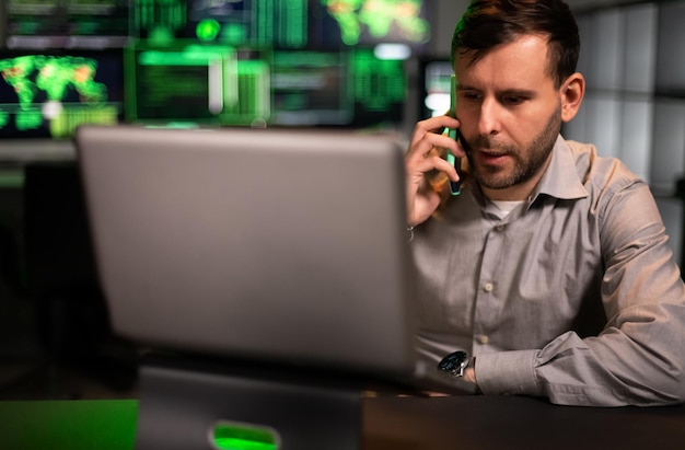 Männlicher Börsenhändler, der im Büro mit Laptop arbeitet und mit dem Kunden am Handy spricht