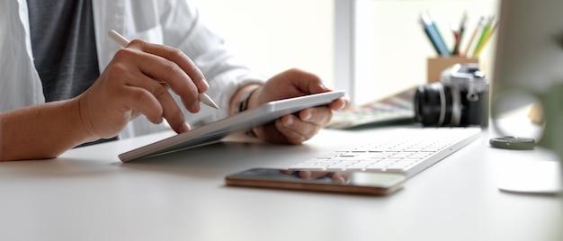 Männlicher Designer, der digitales Tablett mit Stift auf weißem Schreibtisch mit Smartphone, Computergerät und anderen Verbrauchsmaterialien verwendet