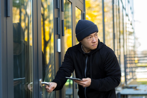 Männlicher Dieb-Hacker versucht, den Alarm im Haus mit einem Virenprogramm zu brechen, verwendet ein Tablet, asiatisch in der Nähe des Hauses