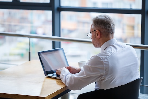 Männlicher Freiberufler arbeitet in einem Café an einem neuen Geschäftsprojekt. Sitzt an einem großen Fenster am Tisch. Schaut mit einer Tasse Kaffee auf einen Laptop-Bildschirm