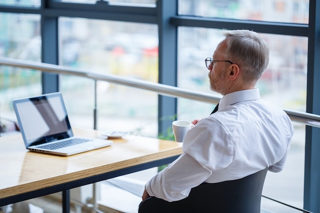 Männlicher Freiberufler arbeitet in einem Café an einem neuen Geschäftsprojekt. Sitzt an einem großen Fenster am Tisch. Schaut mit einer Tasse Kaffee auf einen Laptop-Bildschirm