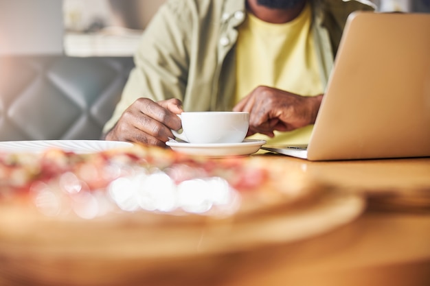 männlicher Freiberufler, der eine Tasse heißes Getränk hält, während er mit einem modernen Laptop im Café am Tisch sitzt?