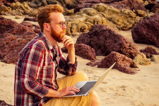 Männlicher Freiberufler mit bärtiger Brille Hipster mit einem Laptop-Fernarbeit am Strand bei Sonnenuntergang.Freiberufler, der in der Nähe der Felsen und des Meeres arbeitet.Geschäftsmann im Urlaub in einem tropischen Wüstenparadies