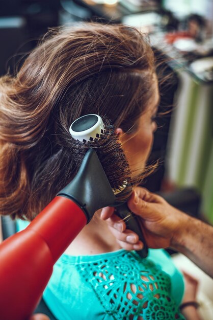 Foto männlicher friseur, der in einem schönheitssalon arbeitet und braunes haar mit runder bürste einer kundin trocknet.