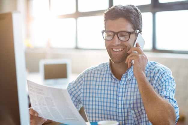 Männlicher Geschäftsleiter, der auf Mobiltelefon beim Lesen der Zeitung spricht