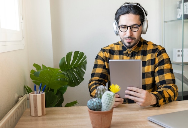 Männlicher Geschäftsmann, der in einem dekorierten Büro sitzt, benutzt ein Tablet, während er mit Kopfhörern Musik hört