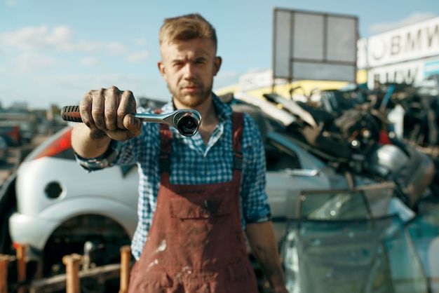 Männlicher Handwerker zeigt Schraubenschlüssel auf Autoschrottplatz. Autoschrott, Fahrzeugschrott, Automüll. Verlassener, beschädigter und zerquetschter Transport, Schrottplatz