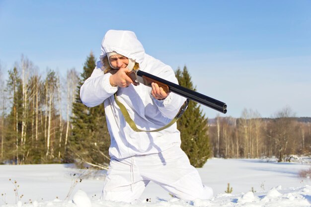 Männlicher Jäger in weißer Tarnung, bewaffnet mit einem Gewehr, das in einem verschneiten Winterwald sitzt