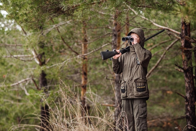 Männlicher Jäger mit Fernglas, bereit zu jagen, Waffe haltend und im Wald spazieren gehend