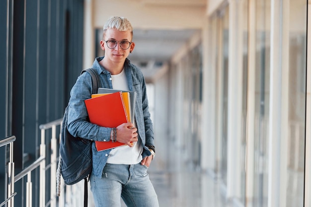 Männlicher junger Student in Jeanskleidung ist im Korridor eines Colleges mit Notizblock in den Händen