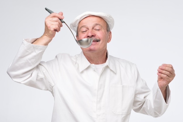 Männlicher Kochkoch in weißer Uniform, der Speisen aus der Pfanne schmeckt