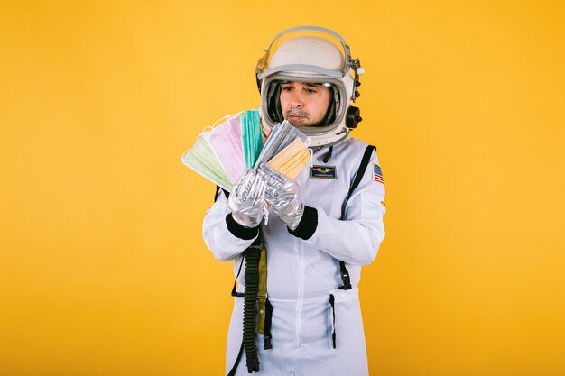 Männlicher Kosmonaut in Raumanzug und Helm, der viele fächerförmige farbige chirurgische Masken auf gelber Wand hält. Covid19 und Viruskonzept