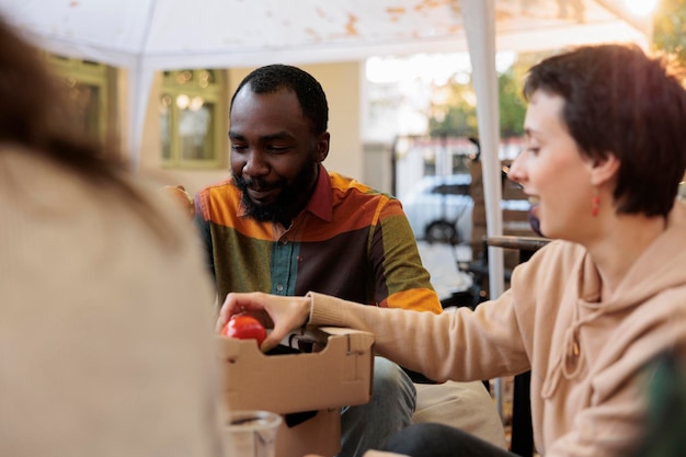 Männlicher Kunde, der im Herbst auf dem Lebensmitteltestfestival Apfel beißt, Kunden, die Spaß mit Besitzern von Bauernmarktständen haben. Junge Leute genießen Bio-Produkte und probieren Obst und Gemüse.