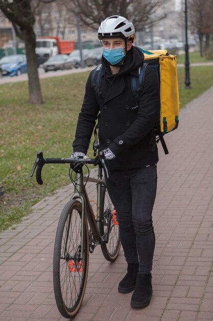 Männlicher Kurier, der medizinische Gesichtsmaske und Thermo-Lieferrucksack trägt und mit Fahrrad geht