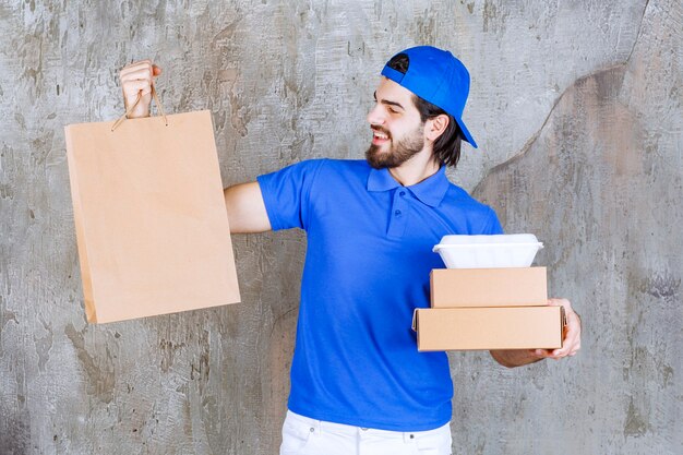 Männlicher Kurier in blauer Uniform mit Papppaketen, einer Einkaufstasche und einer Plastikbox zum Mitnehmen