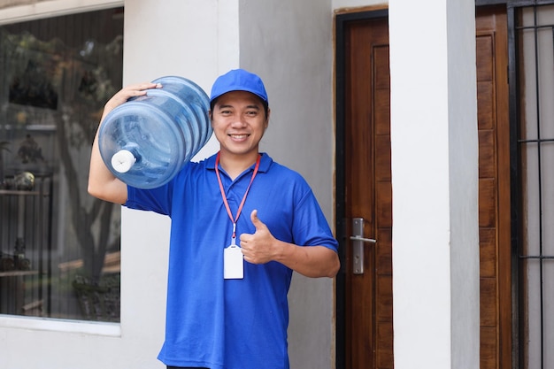 männlicher kurier trägt flasche mit sauberem wasser auf der schulter, bringt auftrag zum kunden und zeigt daumen hoch.