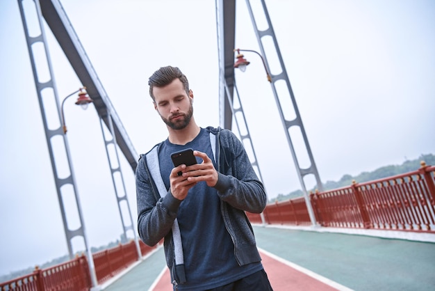 Männlicher Läufer in weißer Windjacke, der bei Sonnenuntergang etwas auf seinem Smartphone tippt