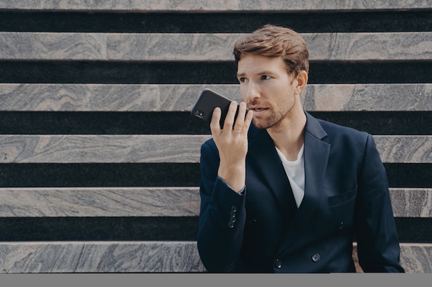 Männlicher leitender Angestellter, der formell gekleidet ist, hält Smartphone in der Nähe von Mundgesprächen auf Freisprecheinrichtung