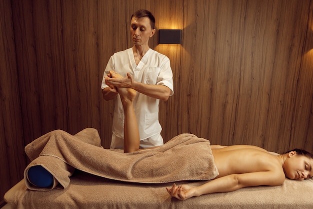 Männlicher Masseur, der der jungen Frau im Handtuch die Beine ausdehnt, professionelle Massage. Massage- und Entspannungstherapie, Körper- und Hautpflege. Attraktive Dame im Spa-Salon