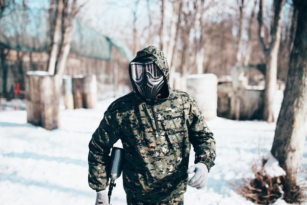 Männlicher Paintballspieler in Schutzmaske und Uniform hält Markierungspistole in Händen, Soldat vor Winterwaldschlacht. Extremsport, militärische Spielausrüstung