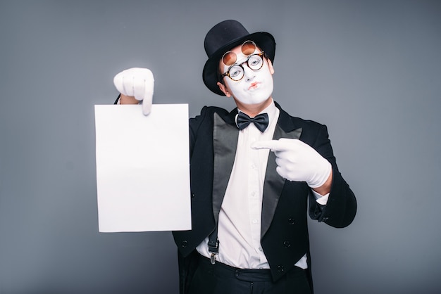 Männlicher Pantomimenschauspieler mit leerem Papierblatt. Pantomime in Anzug, Handschuhen, Brille, Make-up-Maske und Hut.