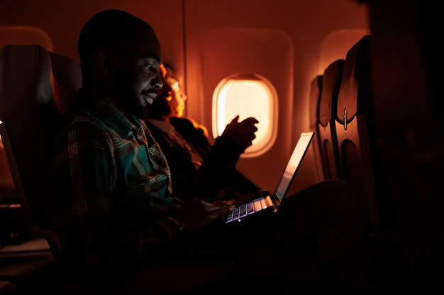 Männlicher Passagier, der auf dem Laptop im Internet surft und im Flugzeug ins Ausland fliegt, mit internationaler Fluggesellschaft, um auf Geschäftsreise zu reisen. Junger Mann, der am Computer arbeitet und während des Sonnenuntergangs unterwegs ist.