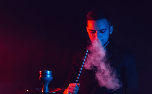 Männlicher Raucher raucht eine Wasserpfeife und stößt eine Rauchwolke vor dem Hintergrund roter und blauer Neonlichter aus