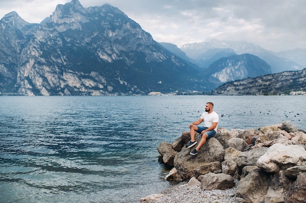 Männlicher Reisender, der auf den Felsen vor dem Hintergrund der Alpen und des Gardasees sitzt. Urlaub in Italien.