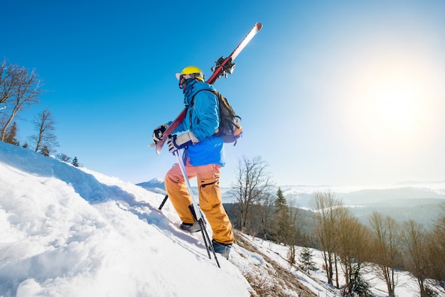 Männlicher Skifahrer, der den Hang in den Bergen hinaufgeht und seine Skier trägt