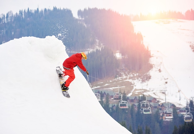 Männlicher Snowboarder, der von der Spitze des schneebedeckten Abhangs mit Snowboard am Winterskigebiet reitet