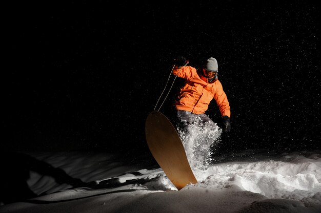 Männlicher Snowboarder in der orange Sportkleidung, die auf dem Snowboard nachts balanciert