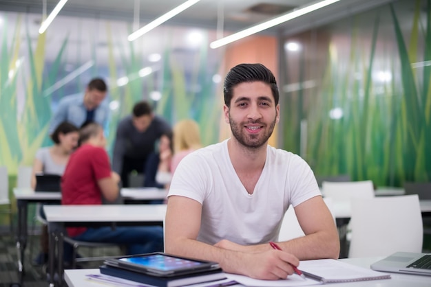 männlicher Student im Klassenzimmer, der Hausaufgaben macht und mit Laptop-Computer lernt, Studentengruppe im Hintergrund