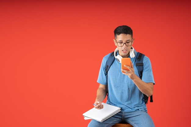 Männlicher Student überrascht, der einen Videoanruf auf seinem Handy macht, während er ein Notizbuch und einen Stift hält