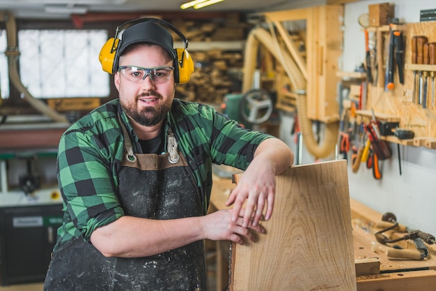 Männlicher Tischler mit Schutzbrille und Kopfhörern lehnt an einer Holzplankenwerkstatt