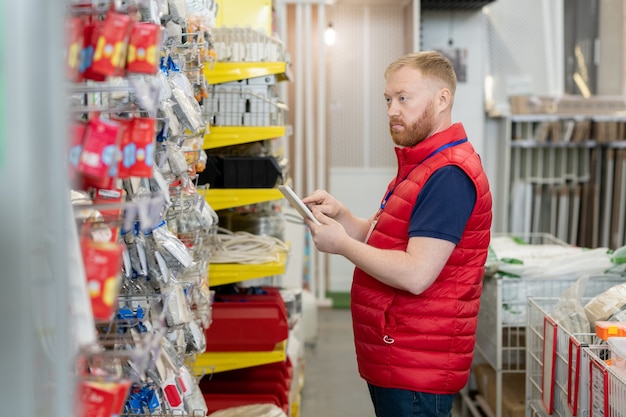 Männlicher Verkäufer in roter Uniform mit Tablet beim Überprüfen von Waren im Hardware-Supermarkt