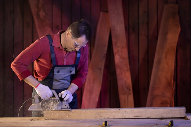 Männlicher Zimmermann mittleren Alters verarbeitet Holzwerkstück mit elektrischem Hobel auf Werkbank in der Werkstatt der Hütte. Tischlerarbeiten an Holz. Arbeiten im Landhaus.