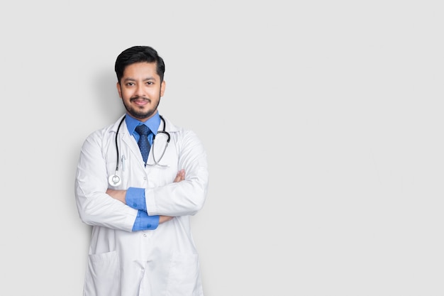 Männliches Doktorporträt, das mit Stethoskop und Armkreuz lokalisiert auf weißer Wand lächelt. Konzept der Krankenversicherung.