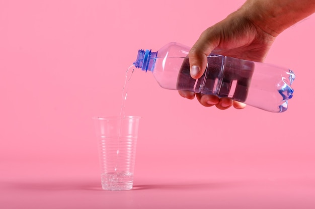 Männliches Gießen des Wassers von der Flasche in Plastikbecher auf rosa Hintergrund