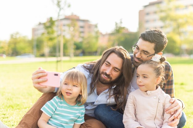Männliches schwules Paar, das ein Selfie mit ihren Kindern macht, die auf dem Gras sitzen
